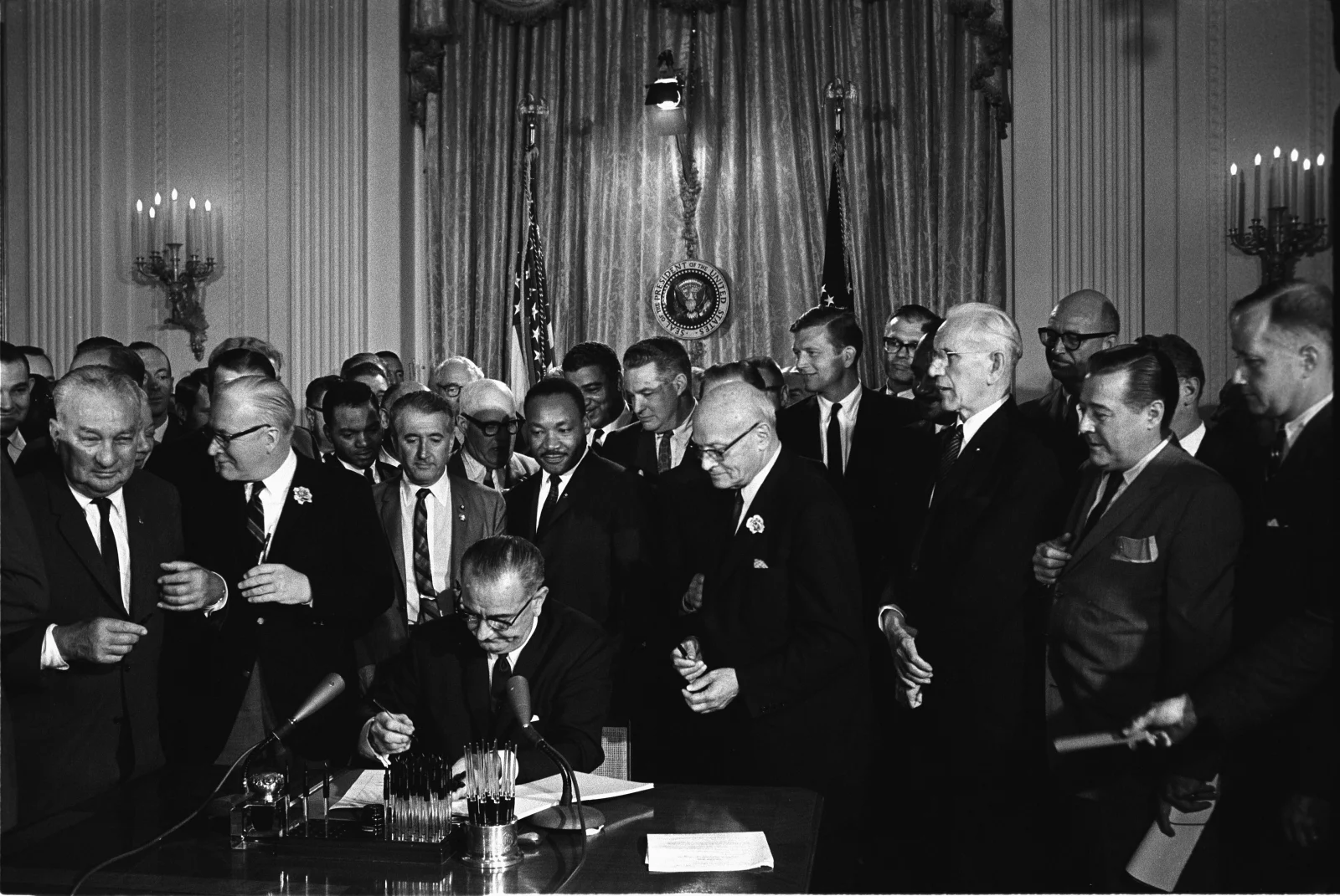 President Lyndon B. Johnson signs the Civil Rights Act at a desk, surrounded by members of Congress and civil rights leaders.
